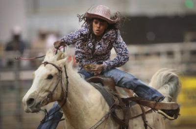 A Black cowgirl in cowboy hat rides a galloping white horse.