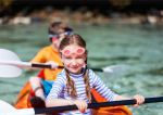 A young girl with swimming goggles on her head paddles an orange double kayak with her brother behind her.