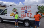 Two female volunteers stand in front of a white van. The van has the "Meet Up & Eat Up" logo on it.