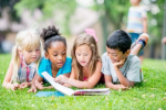 Three girls and one boy lay in the grass as they read a book together.