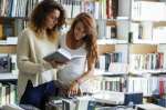 Two women in casual clothing peruse a shelf of books.