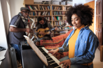 A Black woman playing piano, a Black man playing alto saxophone, and a woman of color playing guitar in a music room.